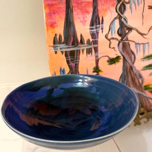 May include: A round, ceramic bowl with a glossy, deep blue glaze featuring darker, swirling indigo and black accents throughout the interior. The exterior rim is a lighter, muted blue, and the bowl has a smooth, curved, and shallow basin shape.