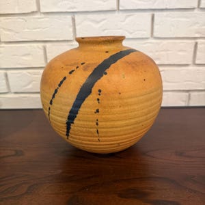 May include: A round, golden-yellow ceramic vase with a narrow neck and a wide, bulbous body. The surface features horizontal ridges along the lower half and a prominent, thick, dark black diagonal brushstroke pattern across the front side.