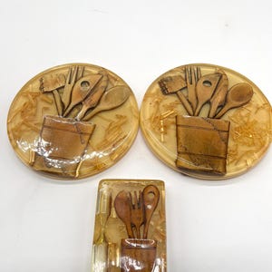 May include: Set of three vintage kitchen-themed coasters. The coasters are made of clear resin with embedded wood shavings and a 3D image of kitchen utensils in a brown container. The coasters are round and rectangular.
