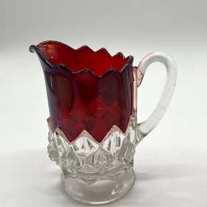 May include: A red glass creamer with a clear glass handle and base. The creamer has a diamond pattern on the base and a scalloped rim.