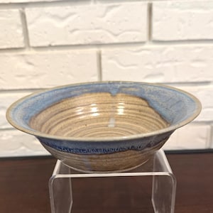 Könnte beinhalten: Eine handgefertigte Keramikschale mit hellbraunem Innenraum und blauem Rand. Die Schale hat ein schlichtes, rustikales Design und wird auf einem transparenten Acrylständer präsentiert. Die Schale eignet sich zum Servieren von Speisen.