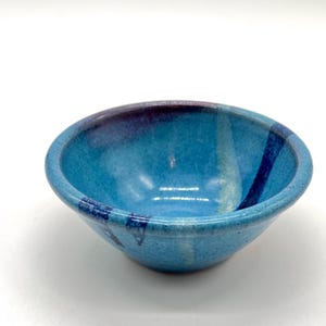 May include: A small, handmade ceramic bowl with a vibrant turquoise glaze. The bowl features streaks of darker blue and purple, creating a unique, artistic design. The bowl is approximately 6 inches in diameter and 3 inches tall.