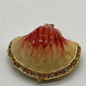 以下が含まれることがあります： 貝殻の形をした装飾的な小物入れ。ボックスはクリーム色のベースに赤オレンジ色のトップがあり、金色のトリムで縁取られています。トリムには、小さなピンクとクリアの宝石がちりばめられています。ユニークな装飾アイテムです。