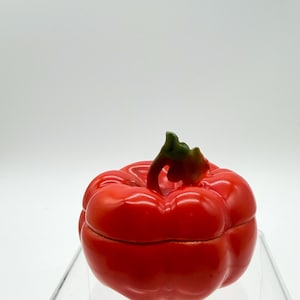 May include: A red, pepper-shaped trinket box with a green stem. The box is glossy and appears to be made of ceramic or a similar material. The lid fits snugly, and the overall design is a realistic representation of a bell pepper.