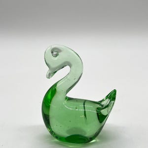 May include: A small, green glass swan figurine. The swan has a white head and neck, and its body is a light green color.