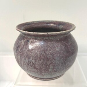 May include: A round, handcrafted ceramic pot with a wide opening. The pot has a speckled, mottled finish in shades of purple, gray, and white. The rim is a lighter shade of purple. The pot is sitting on a clear surface.