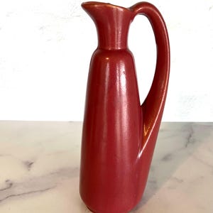 May include: A red ceramic pitcher with a curved handle. The pitcher has a smooth, glossy finish.
