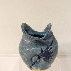 Könnte beinhalten: Eine handgefertigte Keramikvase mit einer einzigartigen, organischen Form. Die Vase ist überwiegend teal mit dunkelblauen Akzenten und einer strukturierten Oberfläche. Sie hat einen kleinen, kreisförmigen Griff und einen ausgestellten Rand. Der Boden hat eine hellbraune Farbe.