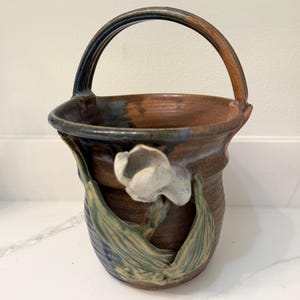 以下が含まれることがあります： 手作りのセラミックバスケット。白い花と緑の葉が彫刻されています。バスケットは茶色のベースに青と茶色のアクセントがあります。ハンドルは濃い青と茶色です。