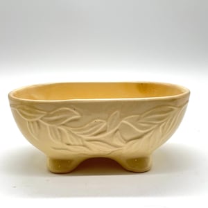 Puede incluir: Un cuenco de cerámica de color crema, de forma ovalada, con tres patas pequeñas. El exterior presenta un diseño de hojas en relieve. El interior del cuenco es de un tono crema ligeramente más oscuro. El cuenco está sobre un fondo blanco.