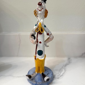 May include: A clown figurine with a white body, decorated with red and green spots, and a yellow collar. The clown has orange hair, a red nose, and a small black hat. The figurine stands on a blue base.