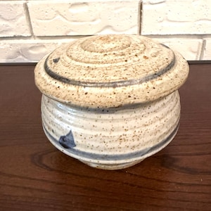 Op de afbeelding: Een keramische botervloot met een deksel. De schaal is gebroken wit met bruine spikkels en een blauwe streep. Het deksel heeft een spiraalvormig ontwerp en een donkerblauwe accent. De schaal staat op een donker houten oppervlak.
