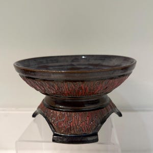 May include: A dark brown ceramic bowl with a textured red and brown base. The bowl has a wide, shallow design and sits on a pedestal base with a textured pattern. The overall aesthetic is rustic and handcrafted.