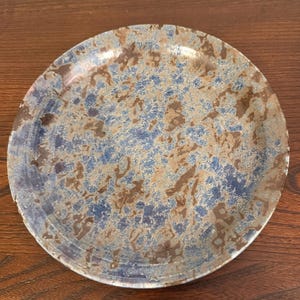May include: A round ceramic plate with a speckled design. The plate features a blend of blue, brown, and tan colors. The rim of the plate has a darker blue hue, and the center has a mottled pattern.
