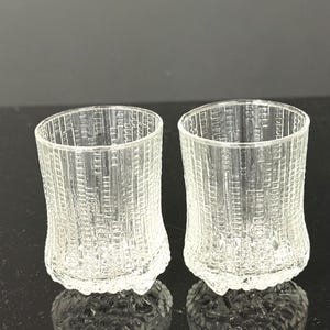 May include: A pair of clear glass tumblers featuring a textured, vertical, and grid-like pattern on the exterior surface. The glasses have a slightly flared rim and a thick, textured base that provides stability and a distinct tactile quality.