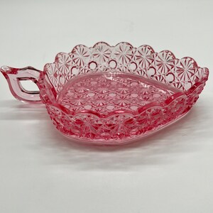May include: A pink glass dish with a scalloped edge and a handle. The dish has a diamond pattern and is perfect for serving small snacks or treats.