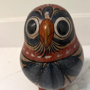 May include: A rounded ceramic owl figurine featuring a hand-painted design with a prominent orange beak, large black eyes, and dark leaf-like patterns on a tan base. The bird has a smooth, glossy finish and stands on two small, sturdy feet.