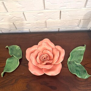 Könnte beinhalten: Ein dreiteiliges Wanddekorationsset aus Keramik, bestehend aus einer großen, pfirsichfarbenen Rose mit geschichteten, geschwungenen Blütenblättern und zwei separaten grünen Blättern mit sichtbarer Blattaderung, alles mit einer glatten Glasur.