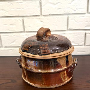 May include: A handcrafted, ceramic lidded pot with a rustic, glazed finish. The pot features a dark brown lid with a loop handle and a matching base with side handles. The color palette includes shades of brown and tan, suggesting a vintage or artisanal style.