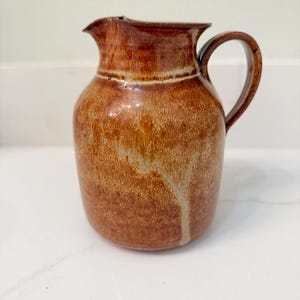May include: A brown ceramic pitcher with a handle and spout. The pitcher has a textured surface with variations in color, ranging from light tan to dark brown. The handle is curved, and the spout is slightly pointed.