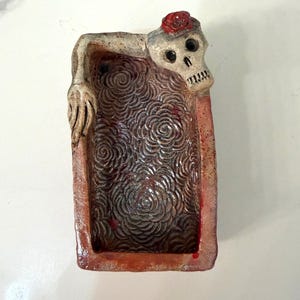 Puede incluir: Un cenicero rectangular de cerámica con temática del Día de Muertos. El cenicero presenta una calavera con una rosa roja en la cabeza y un interior texturizado con un patrón de rosas. Los bordes son de color marrón rojizo.