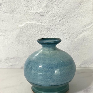 Può includere: Un piccolo vaso rotondo in ceramica con una base larga e un collo stretto. Il vaso è smaltato in tonalità di blu e verde acqua, con una texture macchiettata. Il vaso è alto circa 13 cm.
