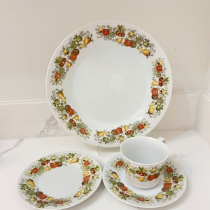 May include: A set of white ceramic dinnerware with a green, yellow and red floral pattern featuring vegetables. The set includes a large dinner plate, a smaller salad plate, and a teacup and saucer.