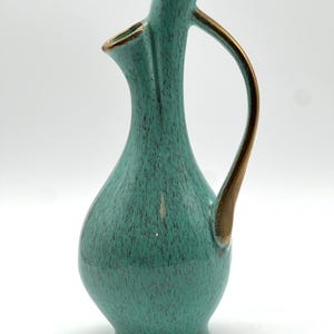 May include: A teal ceramic vase with a unique, asymmetrical design. The vase features a slender neck, a curved handle, and a speckled texture. Gold-colored trim accents the rim and handle, adding a touch of elegance to this decorative piece.