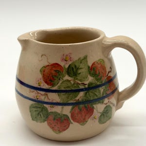May include: A beige ceramic pitcher with a handle and spout. The pitcher is decorated with red strawberries, green leaves, and small pink flowers. Two blue stripes encircle the pitcher. The pitcher has a rustic, handmade appearance.