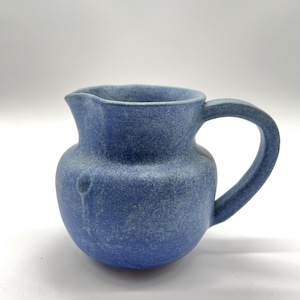 Puede incluir: Una jarra de cerámica azul pequeña con un cuerpo redondeado y un asa curva. La jarra tiene una superficie texturizada y un pico para verter. El color es un azul apagado con motas más claras, lo que le da un aspecto rústico. La jarra es probablemente hecha a mano.