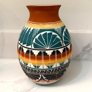 Puede incluir: Un jarrón de cerámica pintado a mano con un cuerpo ancho y un cuello estrecho. El jarrón presenta un borde marrón, patrones geométricos turquesa y blancos, y una banda colorida de naranja, amarillo y negro. El jarrón mide unos 25 cm de alto.