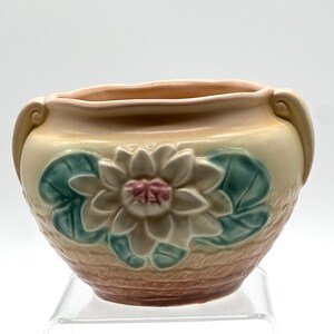 May include: A ceramic bowl with a floral design. The bowl is a light beige color with a pink interior and handles. A white flower with a pink center and green leaves is on the front of the bowl.