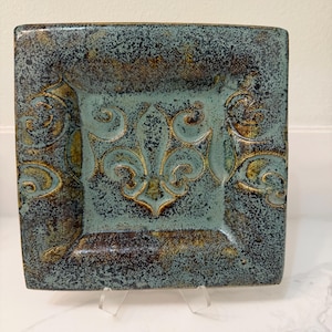 Pode incluir: Prato quadrado de cerâmica com um design de flor de lis. O prato apresenta uma superfície texturizada com um esquema de cores verde-azulado e castanho. O prato é suportado por um suporte transparente.