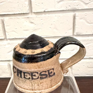 Könnte beinhalten: Ein Keramik-Käsetopf mit Deckel und Griff. Der Topf ist beige mit einem schwarzen Deckel und Griff. Das Wort "CHEESE" steht in schwarzen Buchstaben auf der Seite des Topfes. Der Topf steht auf einer transparenten Oberfläche.