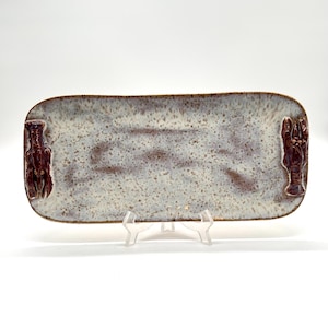 May include: Rectangular ceramic serving platter with a speckled white and brown glaze. The platter has a brown rim and features a raised lobster design at each end. The platter is displayed on a clear plastic stand.