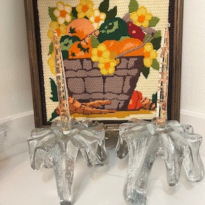 Puede incluir: Dos candelabros de vidrio transparente con una base texturizada en forma de estrella y un tallo delgado. Detrás de ellos hay una imagen enmarcada de punto de aguja de una cesta rebosante de frutas y flores coloridas, en tonos naranja, amarillo y verde.