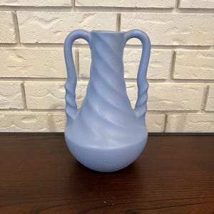 May include: A pale blue vase with two curved handles. The vase features a spiral design and a rounded base. The background is a white brick wall and a dark wooden surface.