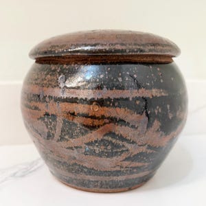 May include: A handcrafted ceramic jar with a lid. The jar is a dark brown color with horizontal bands of lighter brown and speckled accents. The lid is a similar color and texture.