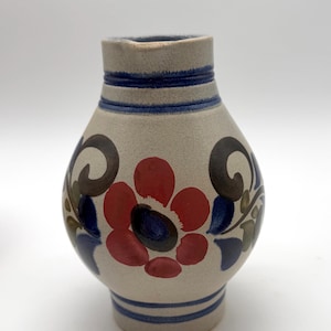 May include: A beige ceramic pitcher with a floral design. The pitcher features a red and blue flower, with blue and green leaves. The neck has two blue stripes, and the body has a bulbous shape.