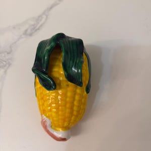 May include: A ceramic corn-on-the-cob figurine with a yellow body and green husk. The figurine has a white base with a pink and red accent. The corn kernels are detailed, and the green husk is folded over the top.