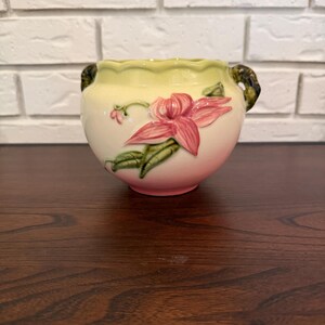 Puede incluir: Una maceta de cerámica con borde festoneado, que presenta un cuerpo de color crema que se degrada hacia una base rosa suave. La maceta está decorada con una flor rosa en relieve y hojas verdes, e incluye dos asas de color verde oscuro.