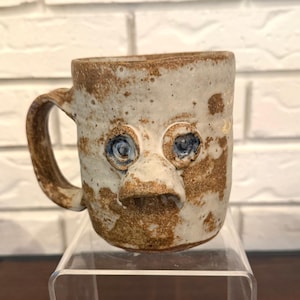 Taza de cerámica firmada con diseño de cara fea y ojos azules.