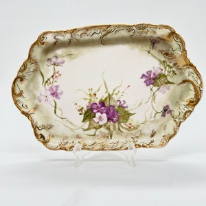 May include: A white porcelain serving tray with a gold rim and a floral design featuring purple and green flowers. The tray has a scalloped edge.