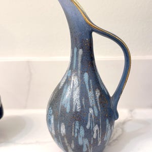 May include: A blue ceramic pitcher with a curved spout and handle. The pitcher has a gold rim and is decorated with white and light blue drips. The pitcher is approximately 10 inches tall.