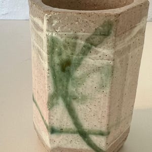 Könnte beinhalten: Eine sechseckige Keramikvase mit hellrosa Glasur und grünen und weißen abstrakten Mustern. Die Vase hat eine raue, strukturierte Oberfläche.