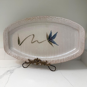 May include: Oval-shaped ceramic serving platter with a floral design. The off-white platter has a textured border and a hand-painted blue flower with green accents. The platter is displayed on a decorative metal stand.