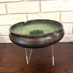 Puede incluir: Un kenzan de cerámica con exterior marrón oscuro e interior verde claro. El kenzan es redondo con una amplia abertura y se apoya en un soporte acrílico transparente. El interior contiene un soporte de alfiler floral verde.