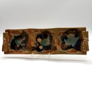 May include: Rectangular ceramic serving dish with three decorative floral designs. The dish has a brown base with orange and blue accents. The ends of the dish have a decorative handle design. The dish is on a clear stand.