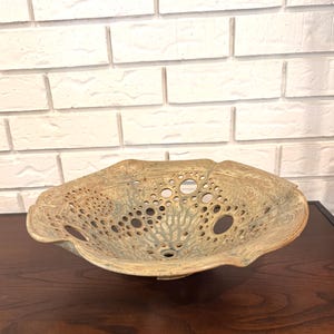 Puede incluir: Un cuenco decorativo de cerámica con un diseño único y orgánico. El cuenco presenta una paleta de colores marrón claro y verde con numerosas perforaciones circulares en toda su superficie. El borde tiene una forma ondulada e irregular, lo que aumenta su atractivo artístico.