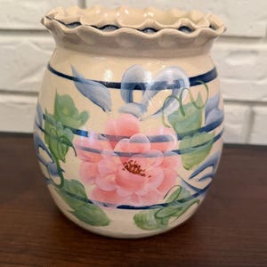 Puede incluir: Un tarro de cerámica de color crema con un borde ondulado y un diseño pintado a mano que presenta una flor grande de color rosa, hojas verdes y finas rayas horizontales azul marino que rodean el cuerpo del recipiente. Acabado liso y brillante.
